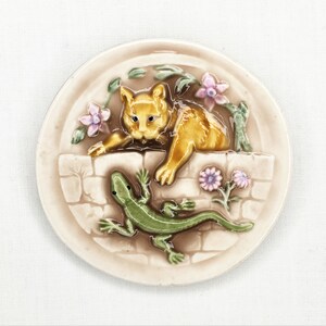 May include: A ceramic plate with a raised design of a cat and a lizard. The cat is orange and white, and the lizard is green. The plate is decorated with flowers and leaves.