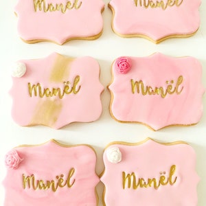 Puede incluir: Seis galletas rosas glaseadas con el nombre "Manël" en letras doradas. Cada galleta tiene un borde decorativo y está cubierta con una pequeña flor comestible. Algunas galletas tienen detalles dorados.