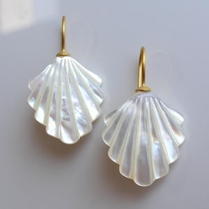 Op de afbeelding: Een paar parelmoerwitte oorbellen in de vorm van een schelp met goudkleurige haken. De oorbellen hebben een waaiervorm met gedetailleerde ribbels, die het licht reflecteren. De sieraden staan tegen een effen, lichte achtergrond.