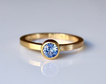 May include: A gold ring featuring a single round, faceted blue gemstone set in a bezel setting. The band has a smooth, matte finish and a consistent width, showcasing a bright, clear blue stone centered on the top of the metal band.