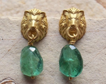 Peut inclure: Une paire de boucles d'oreilles dorées présentant un clou en forme de tête de lion sculptée avec un anneau dans la gueule, d'où pend une pierre précieuse verte translucide et facettée. Le métal est mat et les pierres ont une forme irrégulière.