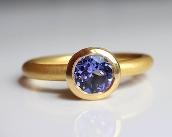 May include: A gold ring featuring a round, bezel-set, light purple gemstone. The ring has a simple, smooth band. The ring is shown on a white surface, reflecting its shape and colour.