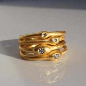 Könnte beinhalten: Ein Stapel von vier goldfarbenen Ringen, jeder mit einer einzigartigen, organischen Form. Jeder Ring hat einen kleinen, klaren Edelstein. Die Ringe werden auf einer weißen Oberfläche mit einem Schatten auf der linken Seite präsentiert.
