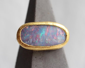 May include: An oval-shaped ring featuring a dark, iridescent opal gemstone with flashes of red, blue, and green light. The stone is set within a textured, matte yellow gold bezel that follows the elongated horizontal shape of the opal.