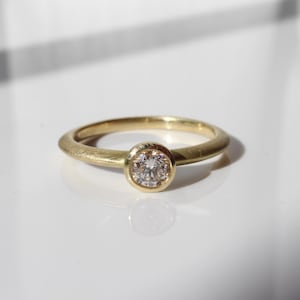 May include: A gold ring with a round, clear gemstone set in a bezel. The ring band is a simple, smooth design. The ring is displayed on a white surface, with shadows adding depth.