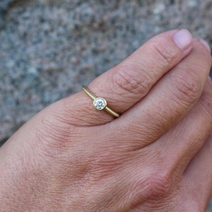 May include: A gold ring with a single, round, clear gemstone set in a bezel. The ring is on a finger, with the hand in the foreground. The background is a blurred, textured surface.