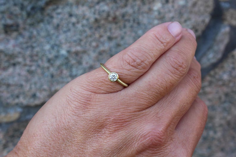 May include: A gold ring with a single, round, clear gemstone set in a bezel. The ring is on a finger, with the hand in the foreground. The background is a blurred, textured surface.