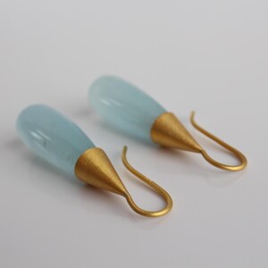 May include: A pair of elegant drop earrings. Each earring features a teardrop-shaped, pale blue gemstone suspended from a gold-colored cone and a curved ear wire. The earrings are displayed against a white background.