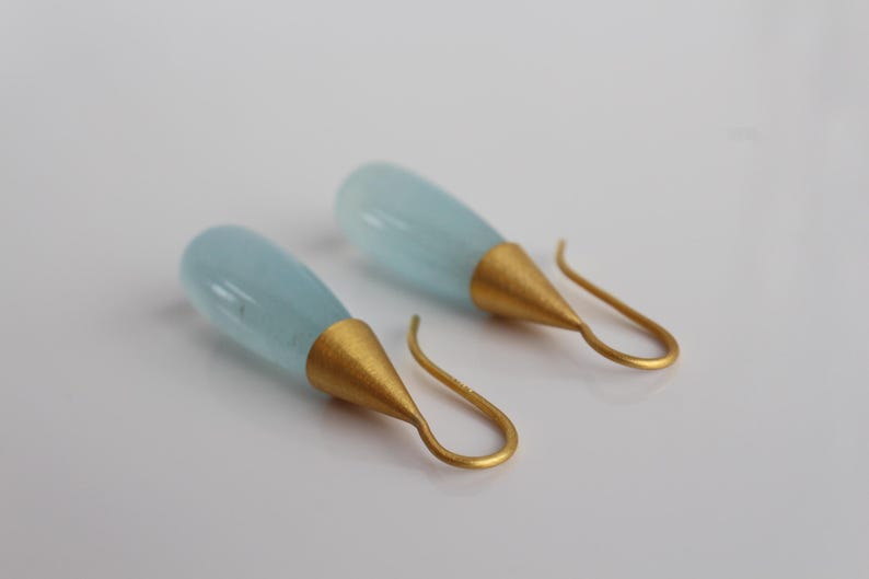 May include: A pair of elegant drop earrings. Each earring features a teardrop-shaped, pale blue gemstone suspended from a gold-colored cone and a curved ear wire. The earrings are displayed against a white background.