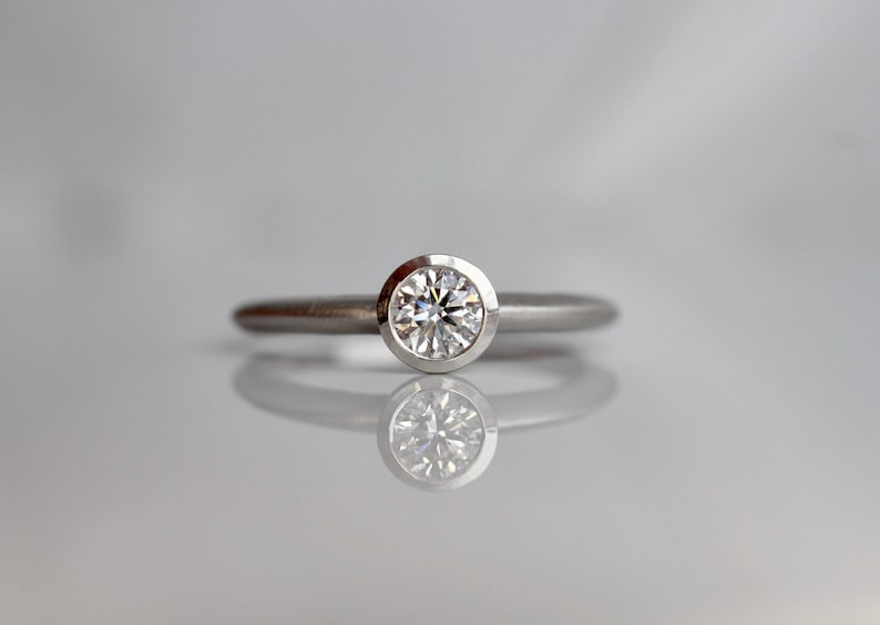 May include: A silver ring with a round, clear diamond set in a bezel. The ring is displayed on a reflective surface, creating a mirrored image of the ring. The band is thin and simple.