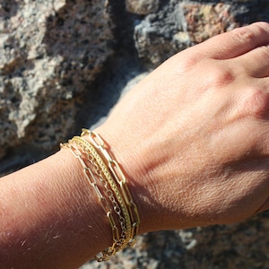 May include: A set of three gold-toned bracelets worn together on a wrist. The set includes one bracelet with elongated rectangular paperclip links, one with a fine twisted rope chain, and one with small, delicate circular links in a layered style.