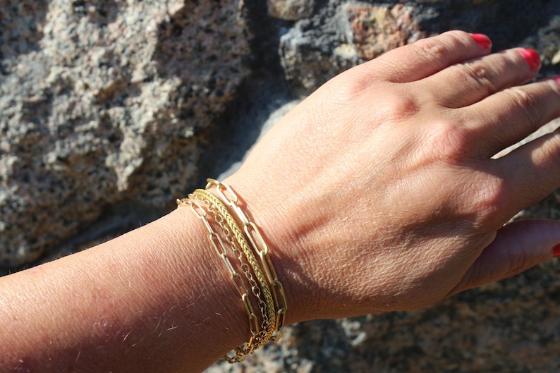 May include: A set of three gold-toned bracelets worn together on a wrist. The set includes one bracelet with elongated rectangular paperclip links, one with a fine twisted rope chain, and one with small, delicate circular links in a layered style.