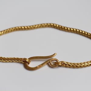 May include: A gold-colored chain bracelet featuring a woven, herringbone-style link pattern. The bracelet is secured with a curved, hook-shaped clasp mechanism that connects to a circular loop at the opposite end of the chain.
