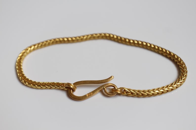 May include: A gold-colored chain bracelet featuring a woven, herringbone-style link pattern. The bracelet is secured with a curved, hook-shaped clasp mechanism that connects to a circular loop at the opposite end of the chain.