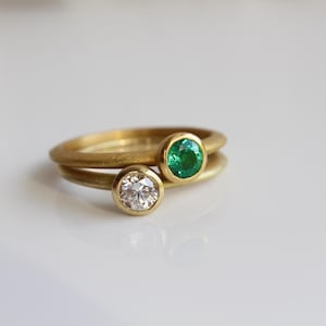 May include: Two gold rings, one with a clear diamond and the other with a green emerald. The rings are stacked, with the emerald ring on top. The rings are set against a white background.