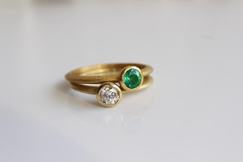 May include: Two gold rings, one with a clear diamond and the other with a green emerald. The rings are stacked, with the emerald ring on top. The rings are set against a white background.