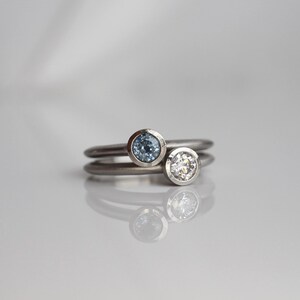 May include: Two silver rings, one with a light blue gemstone and the other with a clear gemstone. The rings are stacked, with the gemstones facing up. The background is a soft, neutral color.