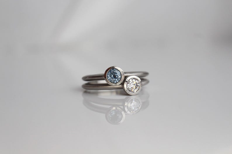 May include: Two silver rings, one with a light blue gemstone and the other with a clear gemstone. The rings are stacked, with the gemstones facing up. The background is a soft, neutral color.