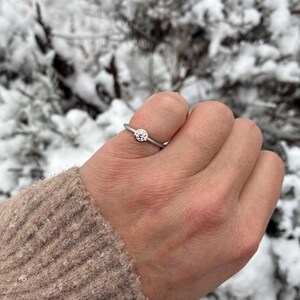 May include: A silver ring with a round, clear gemstone set on a thin band. The ring is on a hand, with a neutral-colored sweater sleeve visible. The background is a snowy, outdoor scene with blurred green foliage.