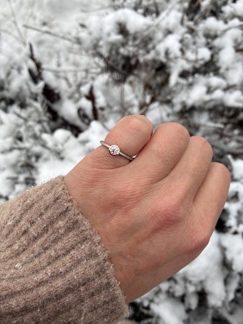 May include: A silver ring with a round, clear gemstone set on a thin band. The ring is on a hand, with a neutral-colored sweater sleeve visible. The background is a snowy, outdoor scene with blurred green foliage.