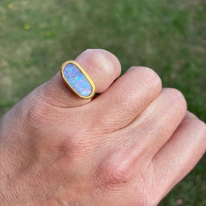 May include: A gold ring featuring an elongated oval-shaped opal gemstone. The opal displays iridescent flashes of blue, pink, and purple colors. The stone is set within a smooth, solid gold bezel setting that encircles the entire perimeter of the gem.