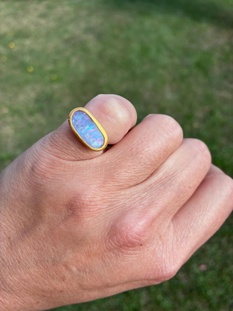 May include: A gold ring featuring an elongated oval-shaped opal gemstone. The opal displays iridescent flashes of blue, pink, and purple colors. The stone is set within a smooth, solid gold bezel setting that encircles the entire perimeter of the gem.