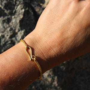 May include: A gold-toned bracelet featuring a fine, woven chain design and a prominent hook-and-eye style clasp. The bracelet is secured around the wrist, showcasing the metallic texture and the distinct curved shape of the central fastening mechanism.