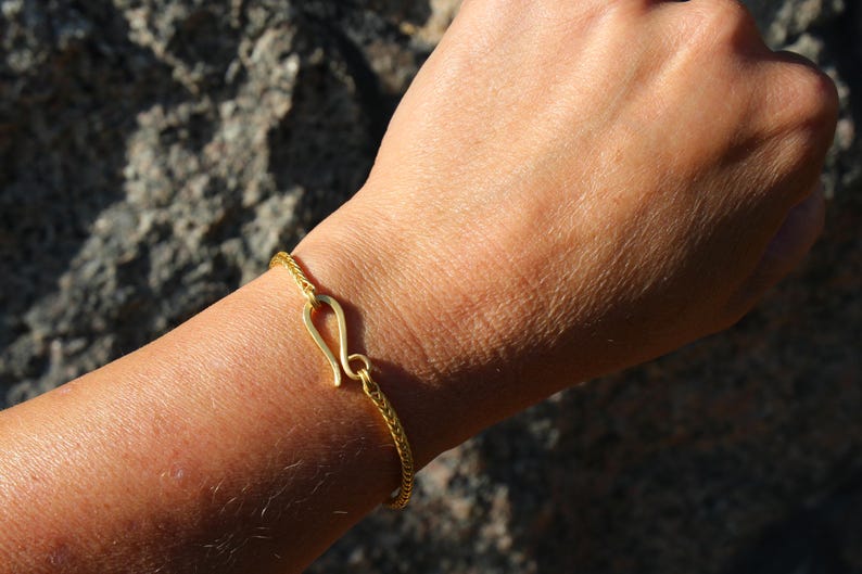 May include: A gold-toned bracelet featuring a fine, woven chain design and a prominent hook-and-eye style clasp. The bracelet is secured around the wrist, showcasing the metallic texture and the distinct curved shape of the central fastening mechanism.