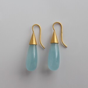 May include: A pair of gold-colored earrings with a teardrop-shaped, light blue gemstone. The earrings have a gold conical top and a curved hook. The background is a plain, light gray color.