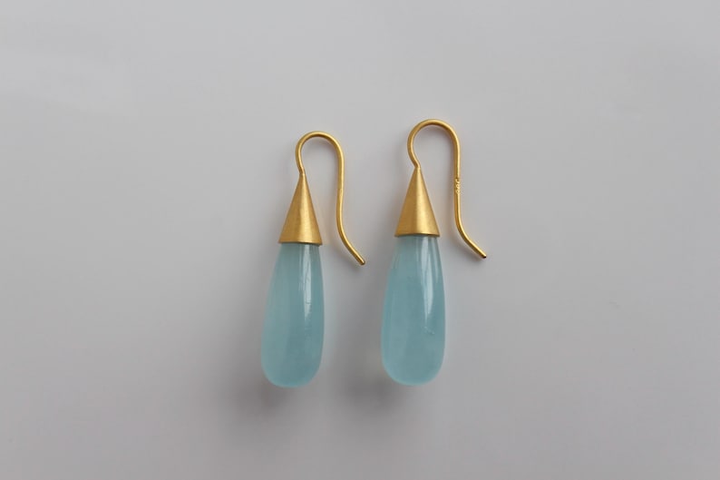 May include: A pair of gold-colored earrings with a teardrop-shaped, light blue gemstone. The earrings have a gold conical top and a curved hook. The background is a plain, light gray color.