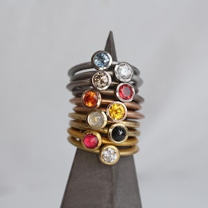 May include: A collection of gemstone rings in various colors, stacked on a dark gray display. The rings feature round gemstones in blue, white, brown, orange, red, yellow, and black. The bands are in silver, copper, and gold tones.