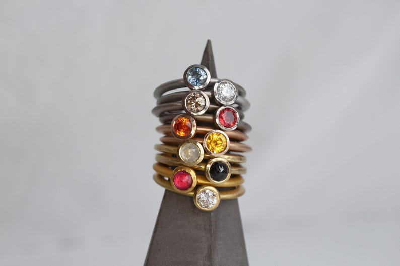 May include: A collection of gemstone rings in various colors, stacked on a dark gray display. The rings feature round gemstones in blue, white, brown, orange, red, yellow, and black. The bands are in silver, copper, and gold tones.
