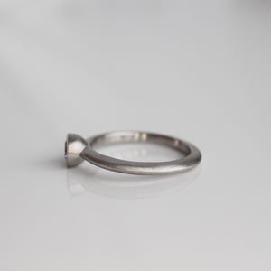 May include: A silver ring with a single, round gemstone. The ring has a simple, elegant design with a smooth, matte finish. The gemstone is set in a bezel setting, and the band is a rounded shape.