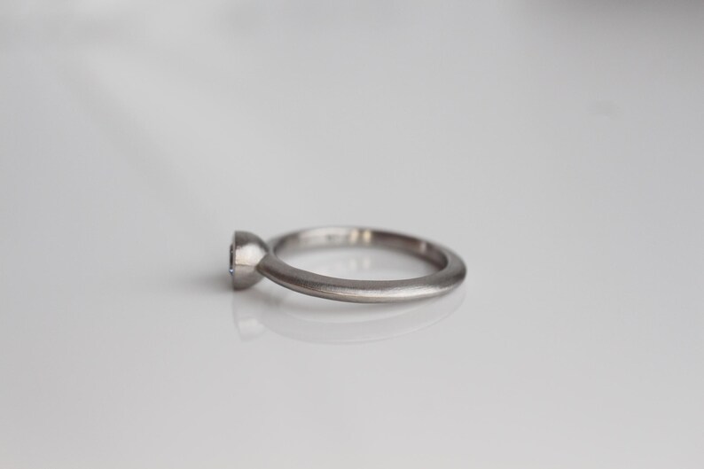 May include: A silver ring with a single, round gemstone. The ring has a simple, elegant design with a smooth, matte finish. The gemstone is set in a bezel setting, and the band is a rounded shape.