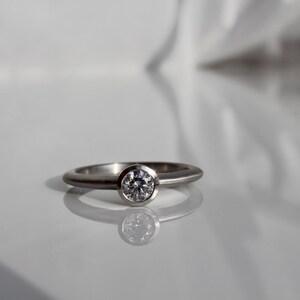 May include: A silver ring with a round, clear gemstone set in a bezel. The ring band is smooth and rounded. The ring is displayed on a reflective white surface.