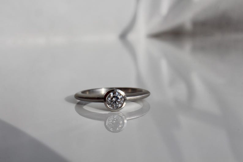 May include: A silver ring with a round, clear gemstone set in a bezel. The ring band is smooth and rounded. The ring is displayed on a reflective white surface.