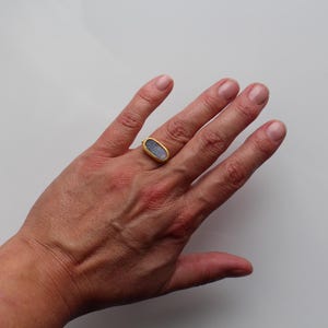 May include: A gold ring featuring an elongated oval-shaped setting with a textured, light blue stone. The metal band has a polished gold finish, and the stone is secured within a simple gold bezel, creating a clean and balanced appearance.