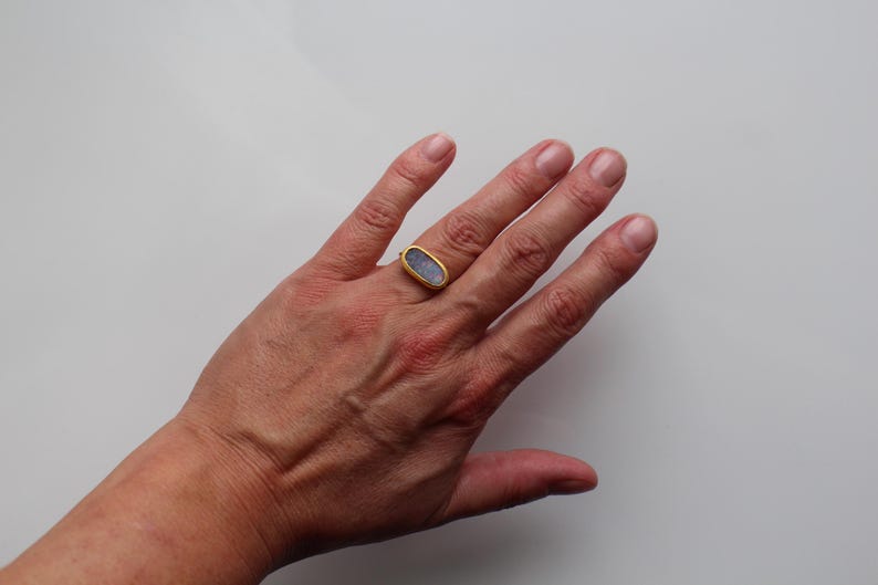 May include: A gold ring featuring an elongated oval-shaped setting with a textured, light blue stone. The metal band has a polished gold finish, and the stone is secured within a simple gold bezel, creating a clean and balanced appearance.