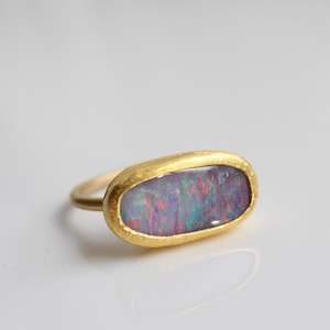 May include: A gold ring featuring an elongated oval-shaped opal gemstone with a textured, matte yellow gold bezel setting. The opal displays iridescent flashes of red, blue, and green color play across its smooth, polished surface.