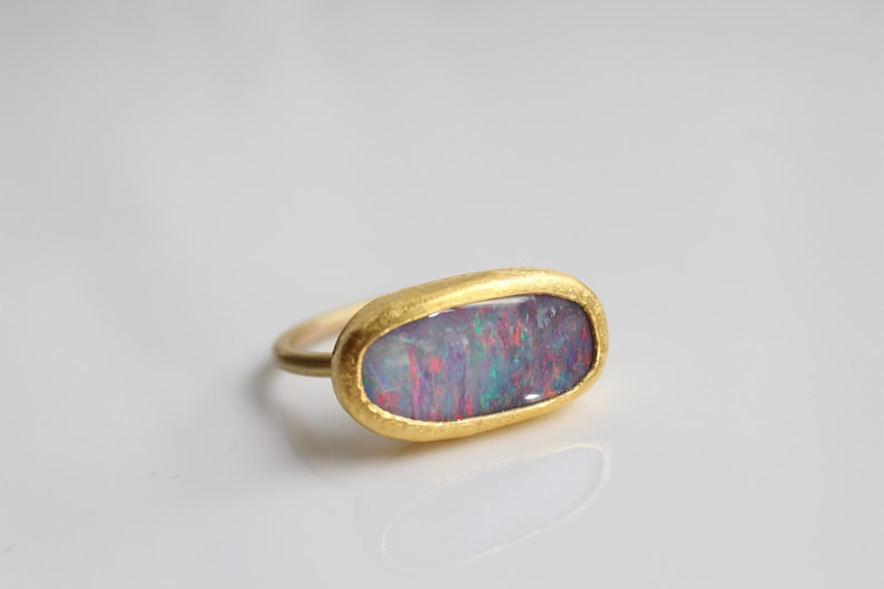 May include: A gold ring featuring an elongated oval-shaped opal gemstone with a textured, matte yellow gold bezel setting. The opal displays iridescent flashes of red, blue, and green color play across its smooth, polished surface.