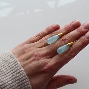 May include: Two aquamarine teardrop earrings with gold-colored caps rest on a hand. The earrings have a light blue, translucent appearance. The gold caps add a touch of elegance to the jewelry.