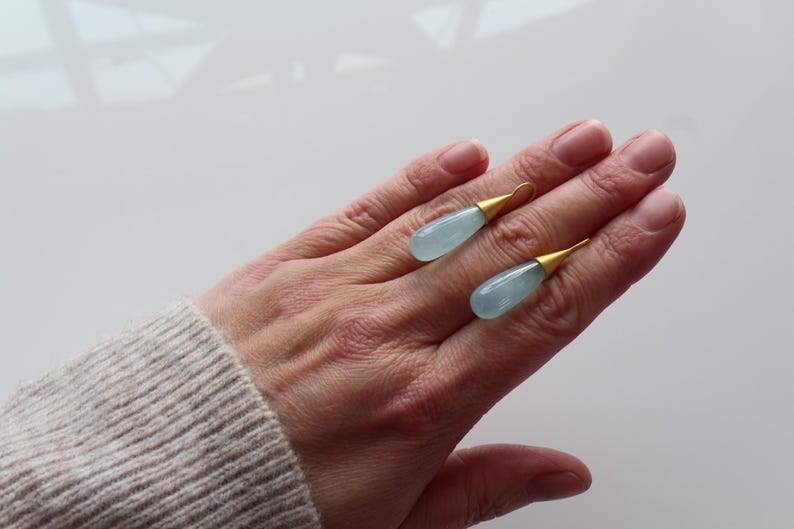 May include: Two aquamarine teardrop earrings with gold-colored caps rest on a hand. The earrings have a light blue, translucent appearance. The gold caps add a touch of elegance to the jewelry.