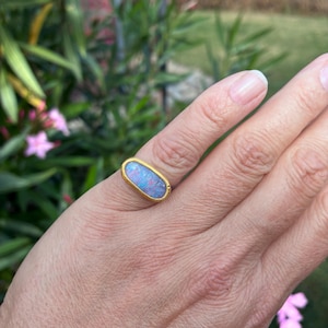 May include: A gold ring featuring an oval-shaped opal gemstone with iridescent flashes of blue and pink. The stone is set in a smooth, polished gold bezel setting that encircles the entire perimeter of the oval opal, creating a simple band design.