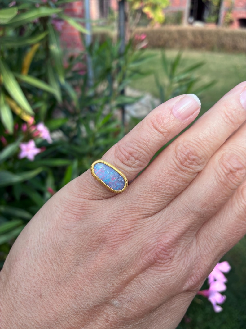 May include: A gold ring featuring an oval-shaped opal gemstone with iridescent flashes of blue and pink. The stone is set in a smooth, polished gold bezel setting that encircles the entire perimeter of the oval opal, creating a simple band design.