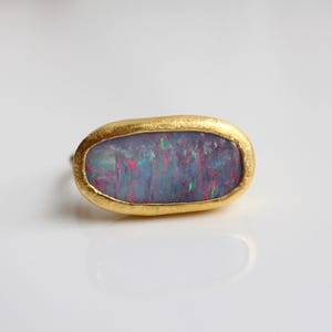 May include: A ring featuring an oval-shaped, dark-toned opal gemstone with iridescent flashes of red, blue, and green. The stone is set within a textured, matte yellow gold bezel that follows the irregular, organic shape of the gemstone.