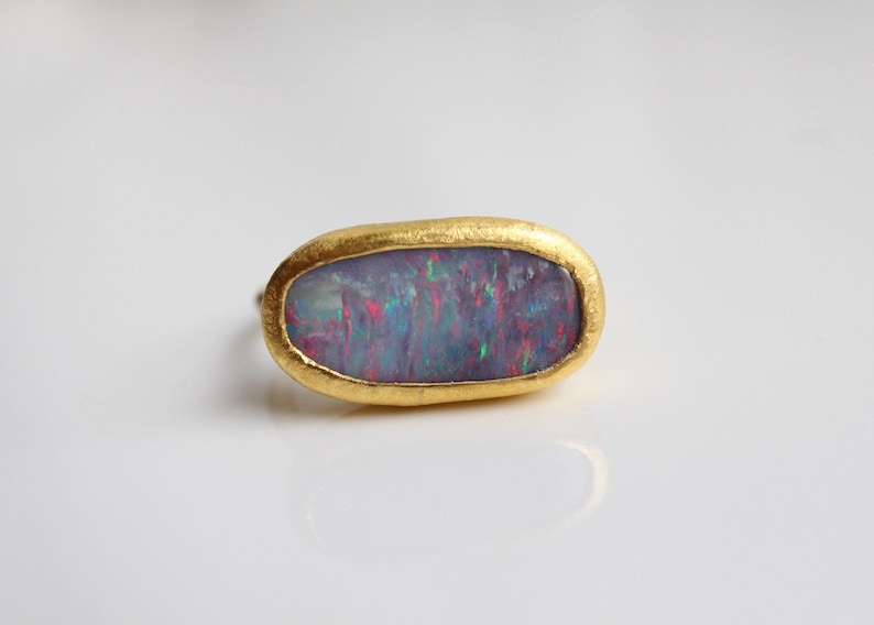 May include: A ring featuring an oval-shaped, dark-toned opal gemstone with iridescent flashes of red, blue, and green. The stone is set within a textured, matte yellow gold bezel that follows the irregular, organic shape of the gemstone.