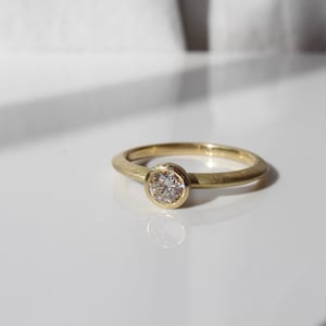 May include: A gold ring with a round, bezel-set diamond. The ring has a simple, smooth band and a bright, polished finish. The diamond is clear and sparkling, set in a circular gold bezel.