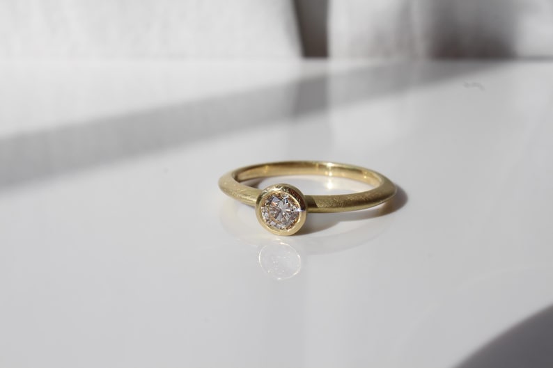 May include: A gold ring with a round, bezel-set diamond. The ring has a simple, smooth band and a bright, polished finish. The diamond is clear and sparkling, set in a circular gold bezel.