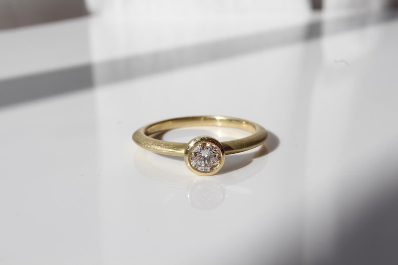 May include: A gold ring with a round, clear gemstone set in a bezel. The ring band is a simple, smooth design. The ring is displayed on a white surface, with shadows adding depth.
