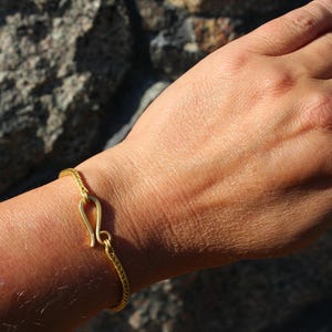 May include: A gold-toned bracelet featuring a fine, textured chain and a prominent hook-and-eye style clasp mechanism. The bracelet is crafted with a matte finish and displays a consistent, intricate woven pattern throughout the entire length of the band.